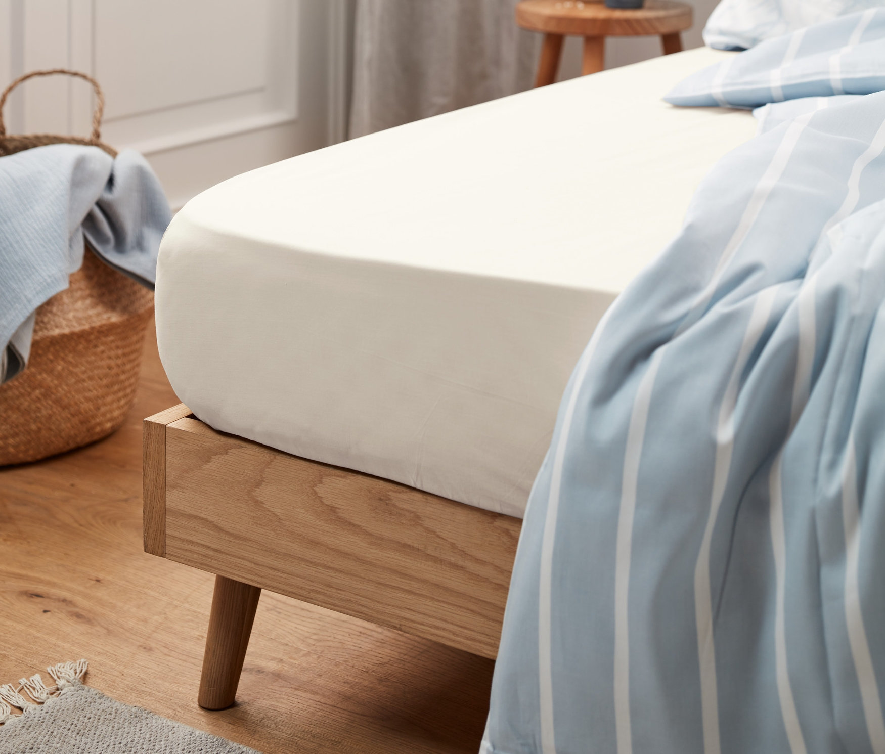 Egy bézs színű, normál méretű jersey lepedő van egy fa keretes ágyra húzva. A háttérben egy kosár kék anyaggal.