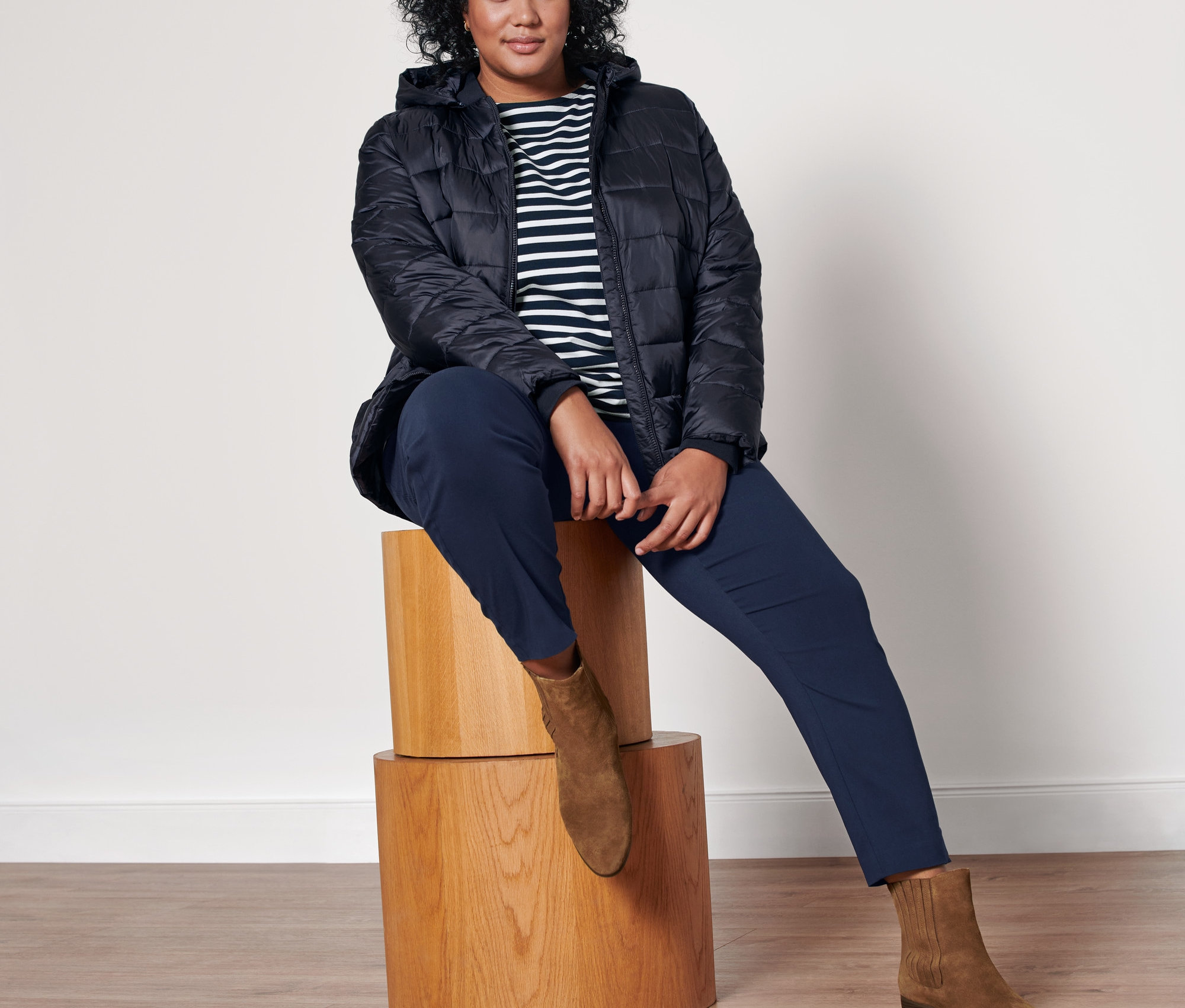 Nő ül egy fa zsámolyon, kék csíkos hosszú ujjú felsőt, sötétkék sztreccsnadrágot, velúrbőr csizmát és sötétkék, kapucnis steppelt kabátot visel.