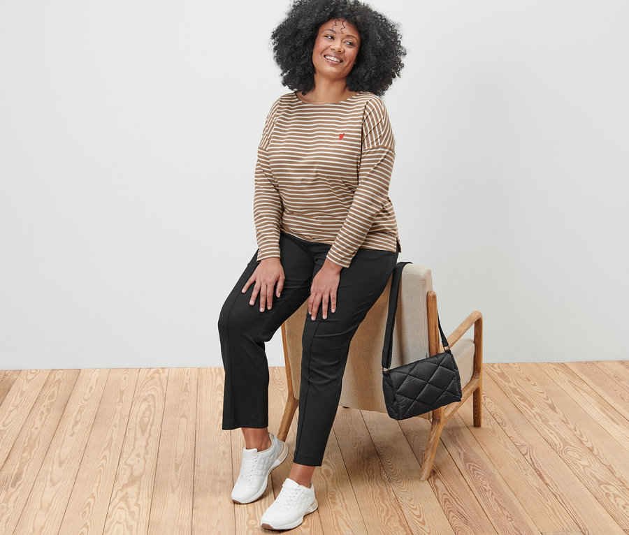 Nő ül egy széken, csíkos hosszú ujjú felsőt, fekete női szabadidőnadrágot és fehér tornacipőt visel. Mellette fekete steppelt oldaltáska látható.