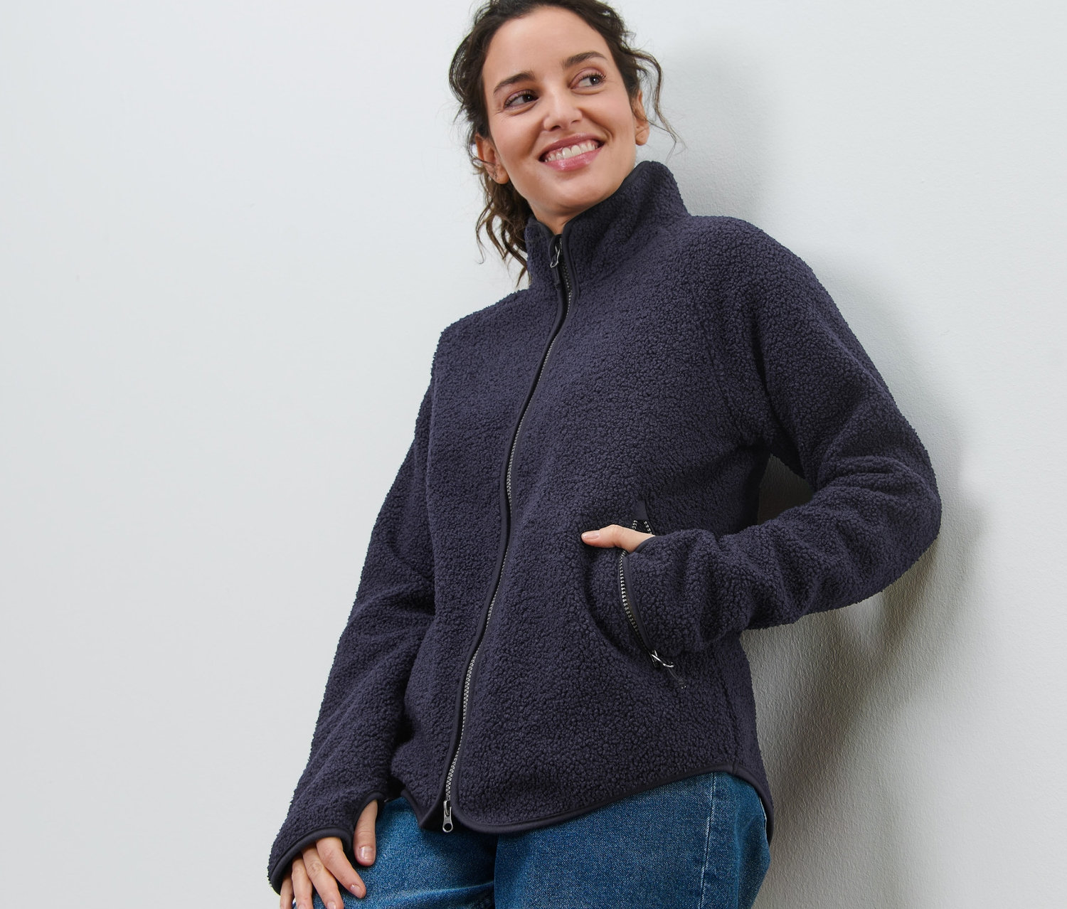 Mosolygó nő a falnak dőlve tengerészkék plüss polár felsőt és farmert visel.