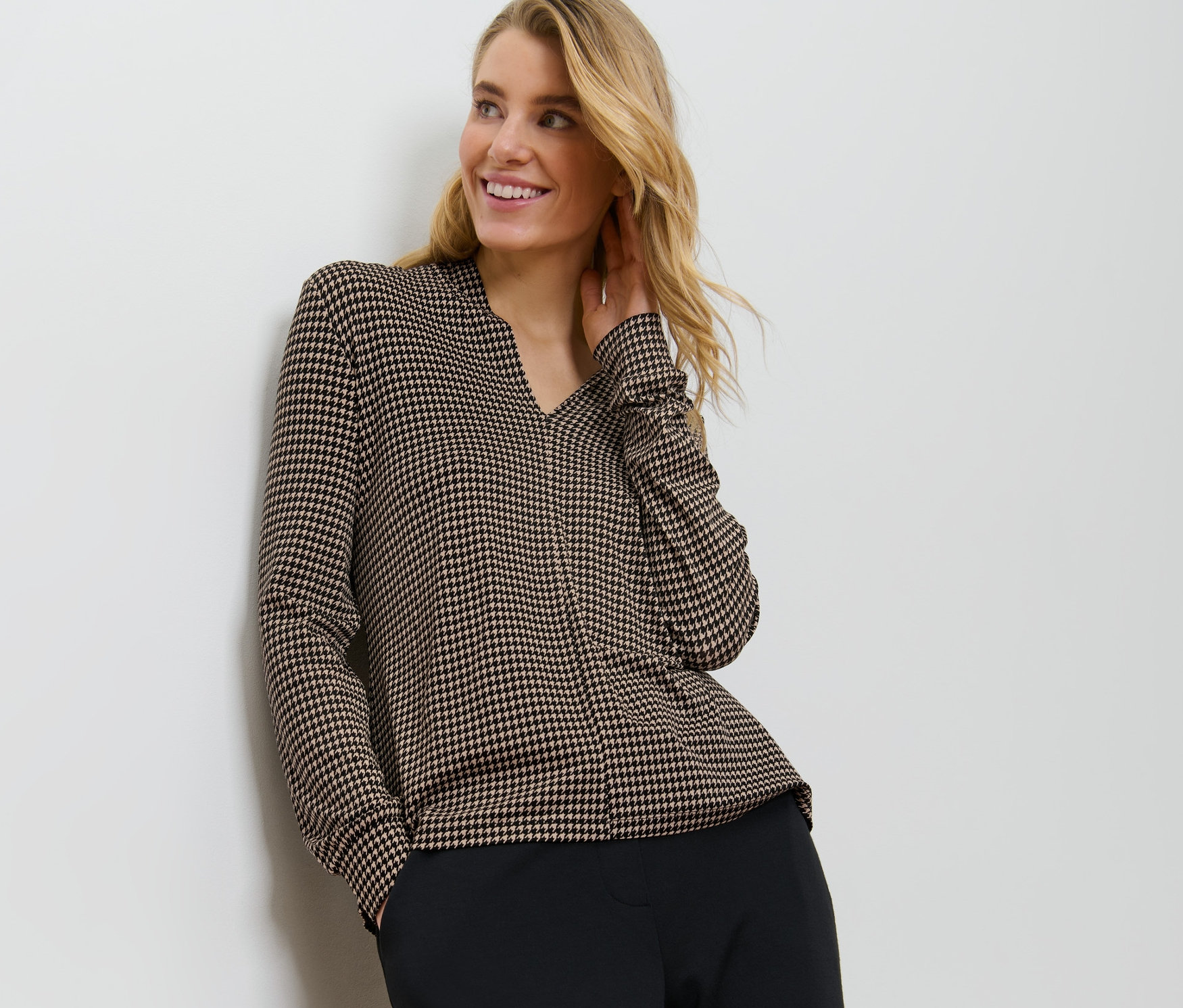 Egy szőke nő a falnak dőlve nyomott mintás pólóblúzt visel V-nyakkivágással.