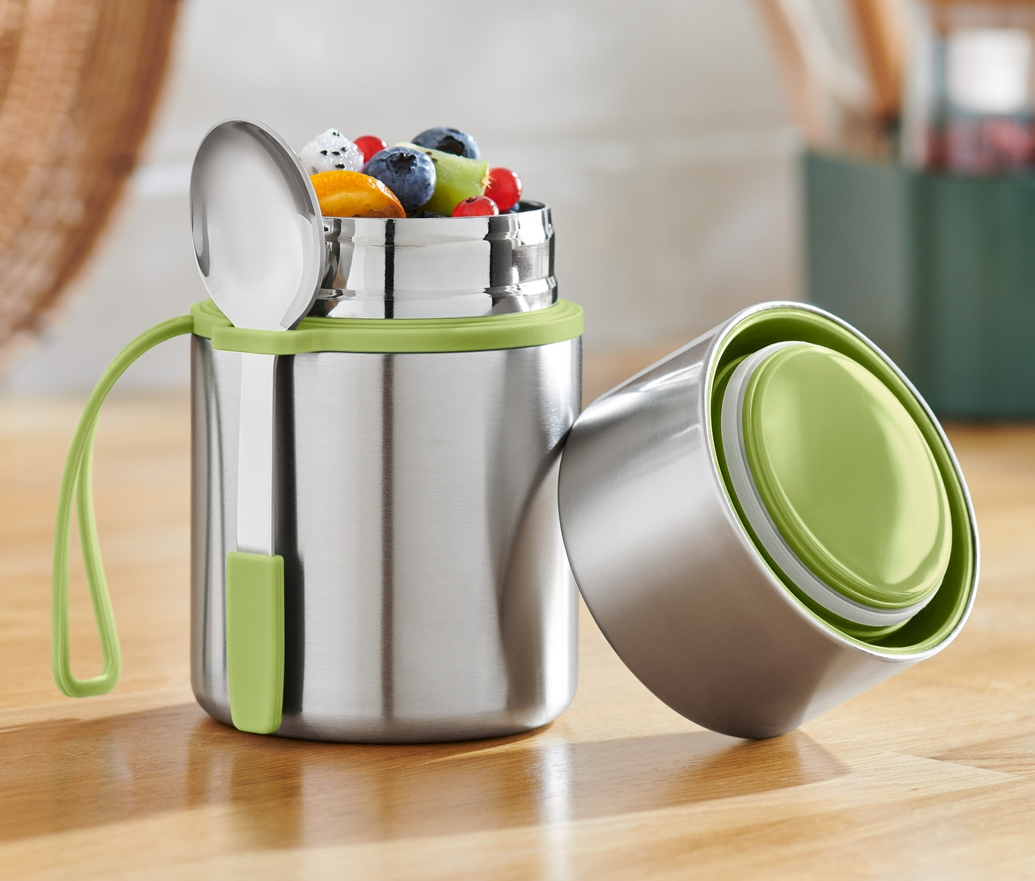Rozsdamentes acél ételhordó termosz gyümölccsel és kanállal egy fa asztalon.