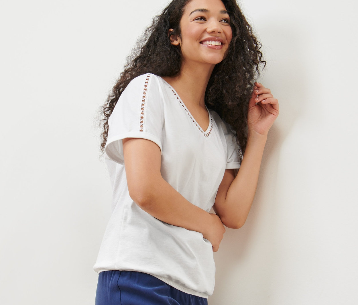 Egy mosolygó, hosszú, göndör hajú nő fehér, horgolt díszítésű blúzt és kék nadrágot visel.