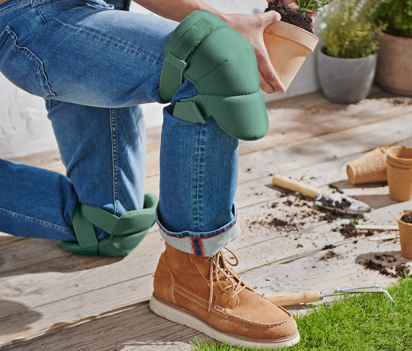 Egy térdelő személy kerti térdkímélő alátéttel egy virágcserepet tart.