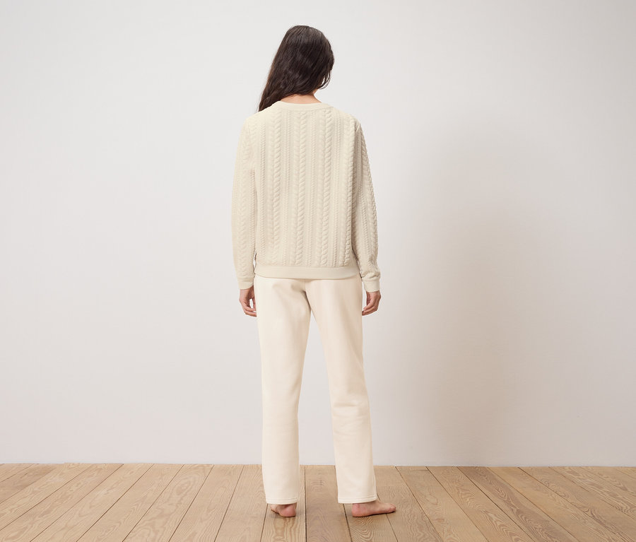 Egy nő áll háttal a kamerának, krémszínű pulóvert és nadrágot visel.