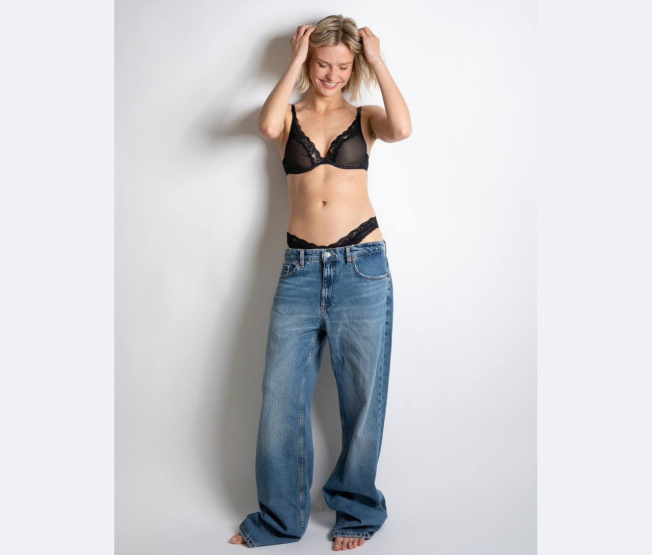 Egy nő mosolyog és a fején tartja a kezét, fekete csipkés melltartót és tangát, valamint hatalmas farmert visel.