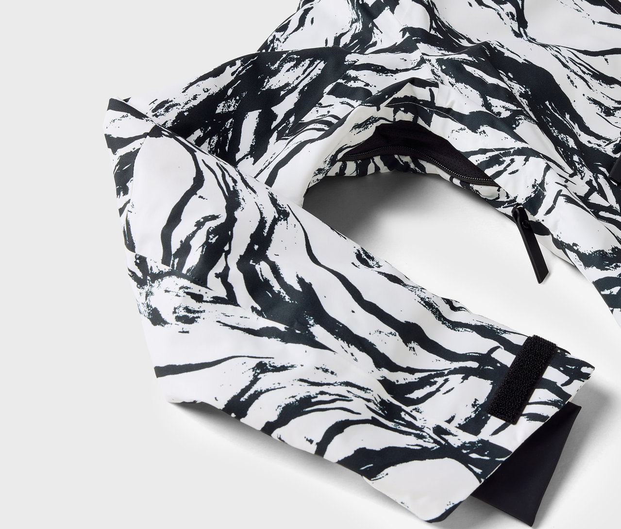 Egy fehér, fekete mintás kabát részlete, látható az ujja és a cipzár.