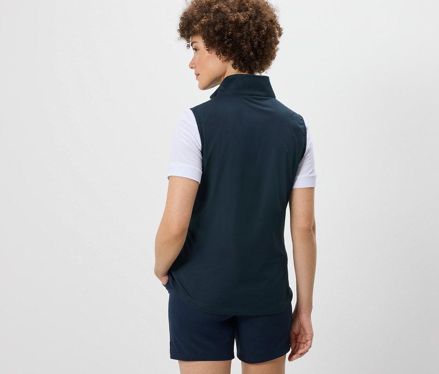 Hátulról látható egy afro frizurás nő, aki softshell mellényt és rövidnadrágot visel.