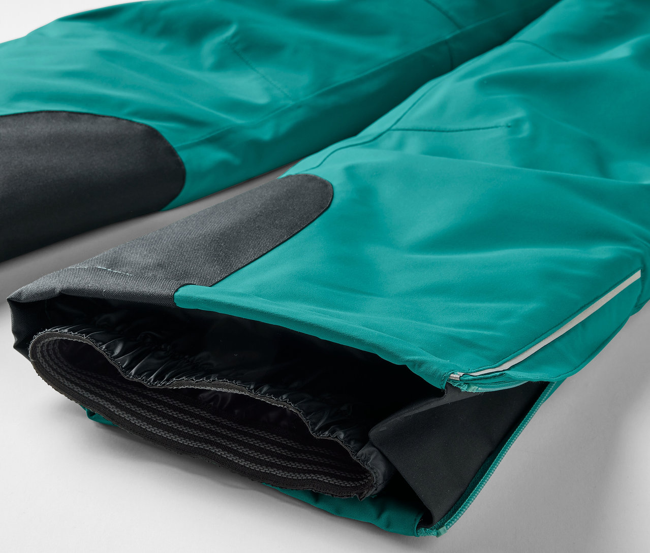 Egy türkiz színű nadrág közeli képe, fekete térdfoltokkal és egy szétnyitott szárral, ami fekete bélést mutat.