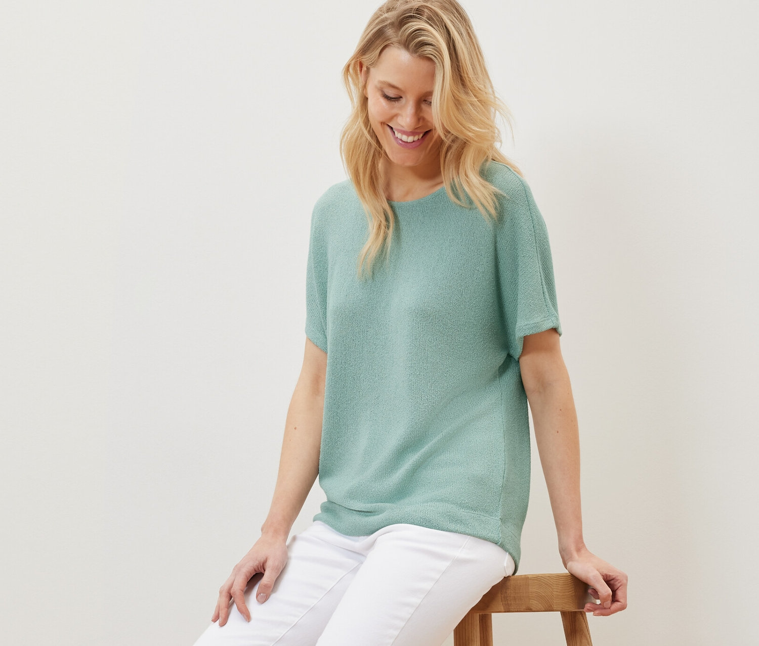 Szőke nő ül egy fa székleten, zsályaszínű strukturált felsőt és fehér nadrágot visel.