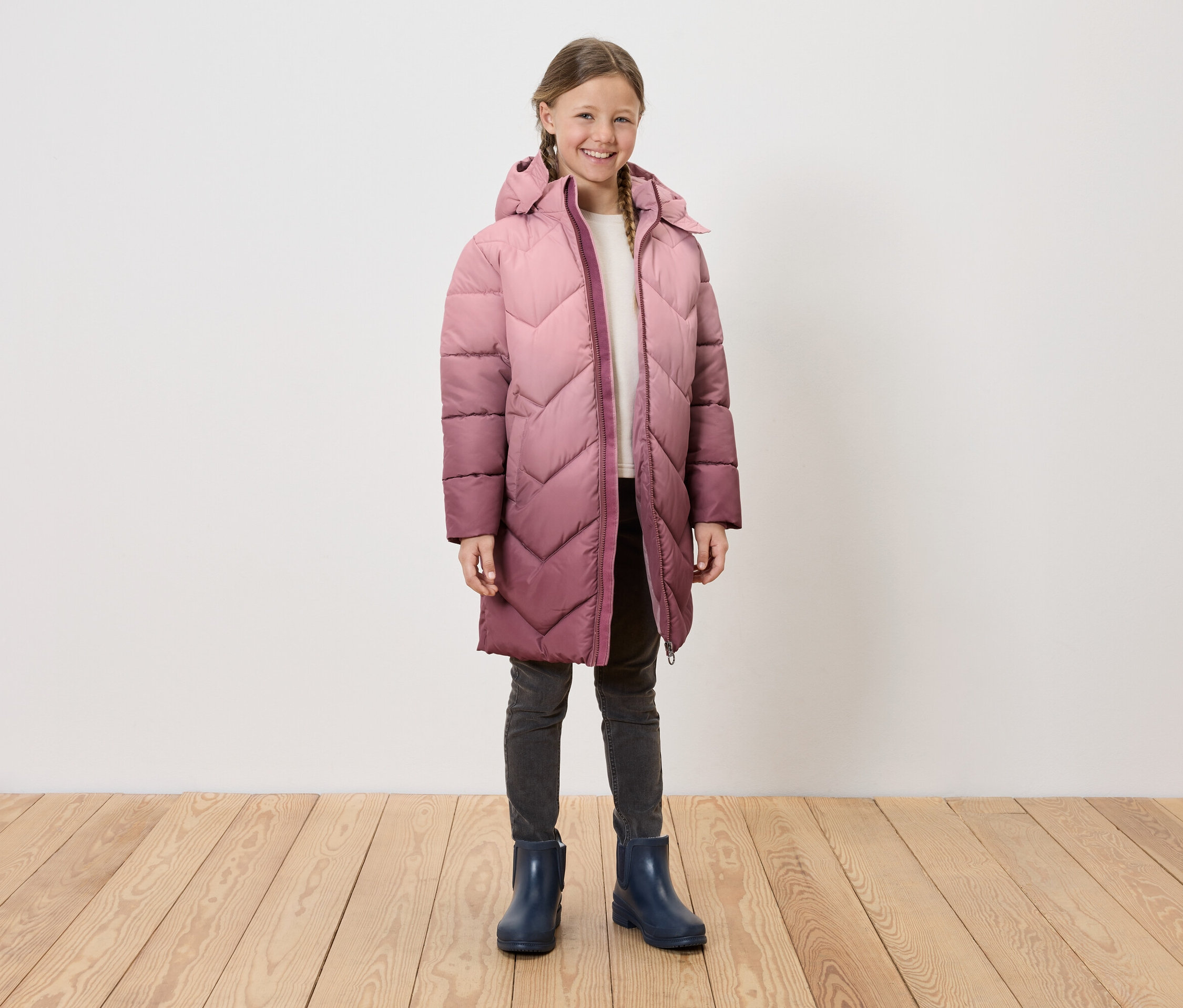 Lány hosszú, steppelt, kapucnis kabátban, szürke farmerban és kék gumicsizmában áll a fapadlón egy fehér fal előtt.