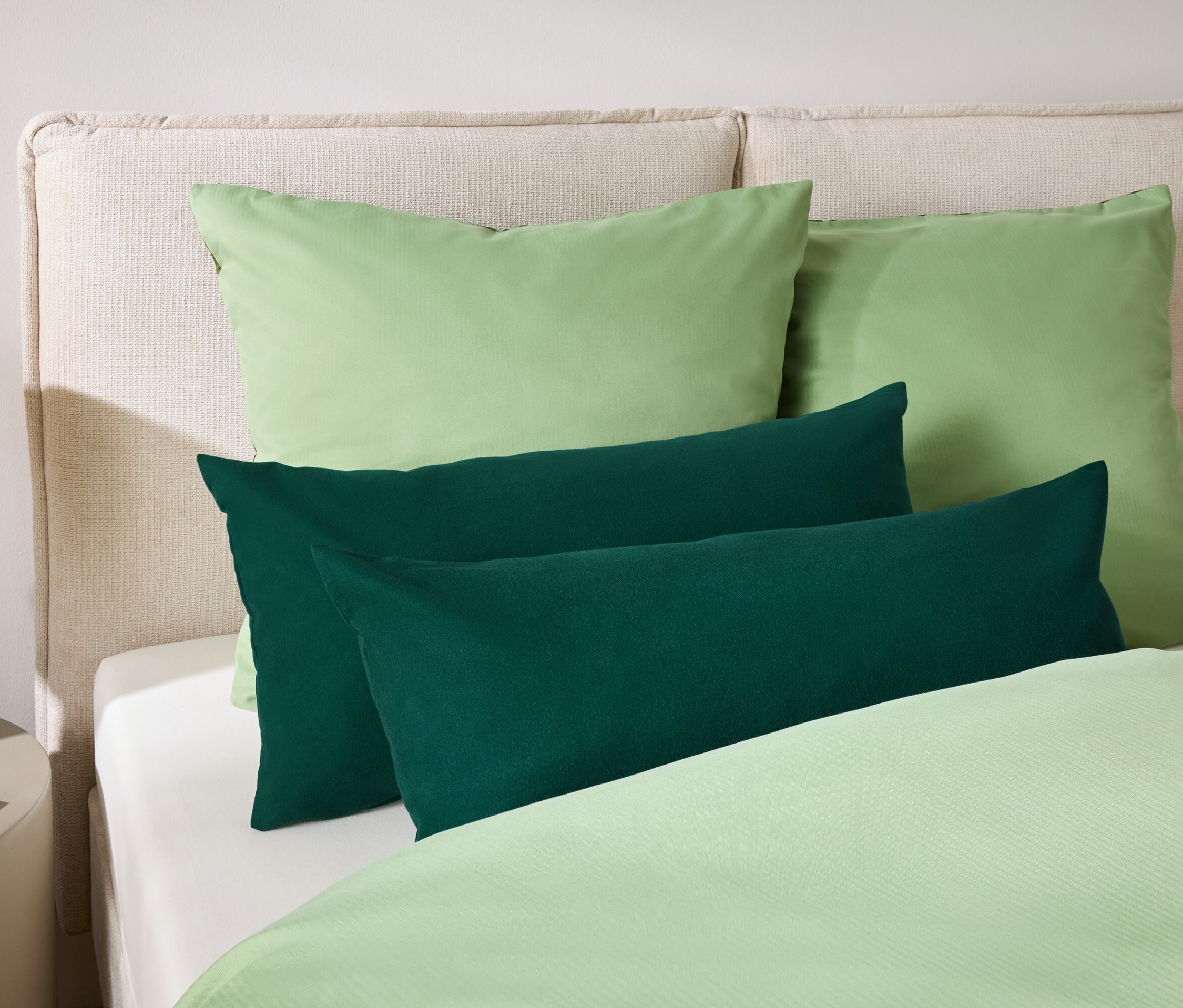 Két zöld párna és két sötétzöld, hosszúkás párna fekszik egy krémszínű fejtámlával és világoszöld ágyneművel ellátott ágyon.
