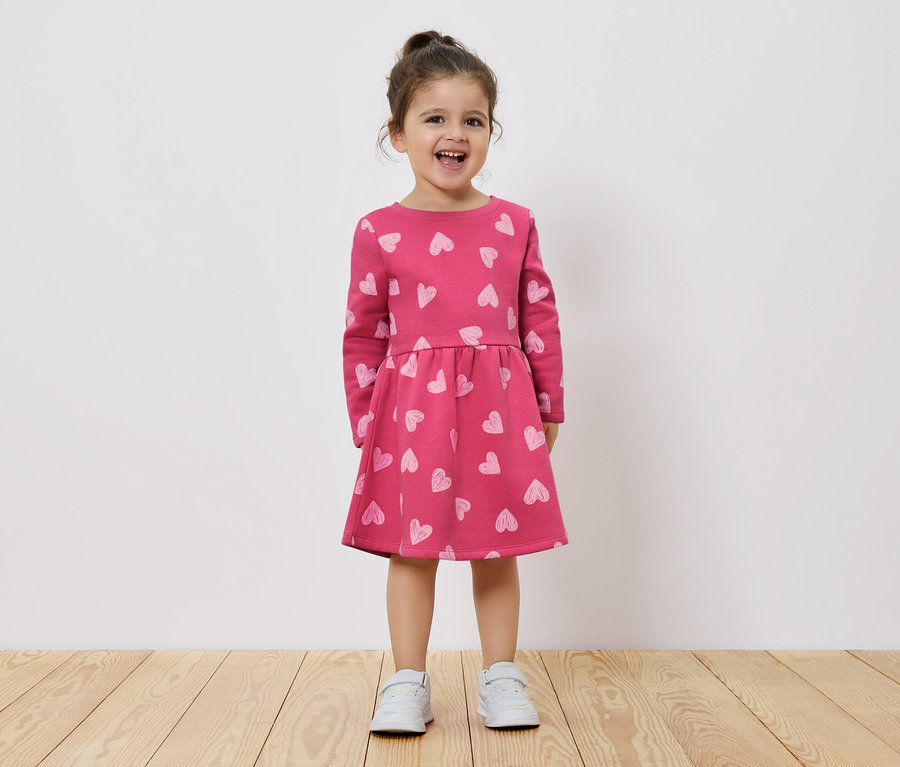 Egy mosolygó kislány áll egy fapadlón, és rózsaszín, hosszú ujjú, szívecskés mintájú ruhát visel.