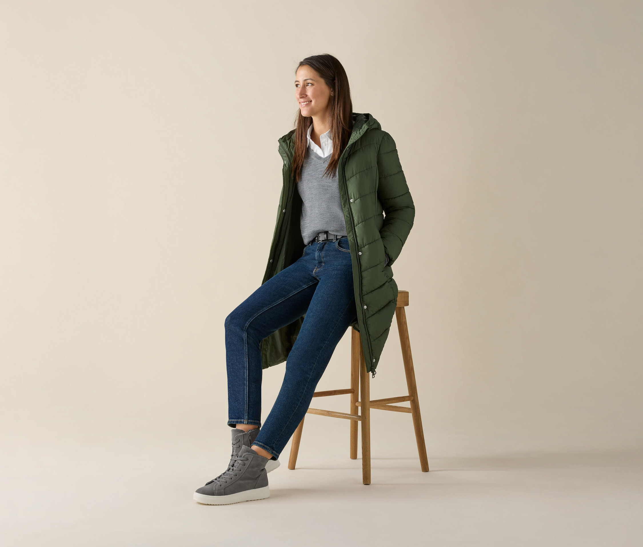 Egy nő ül egy széken, szürke finom kötésű merinó pulóvert, farmert és zöld kabátot visel.
