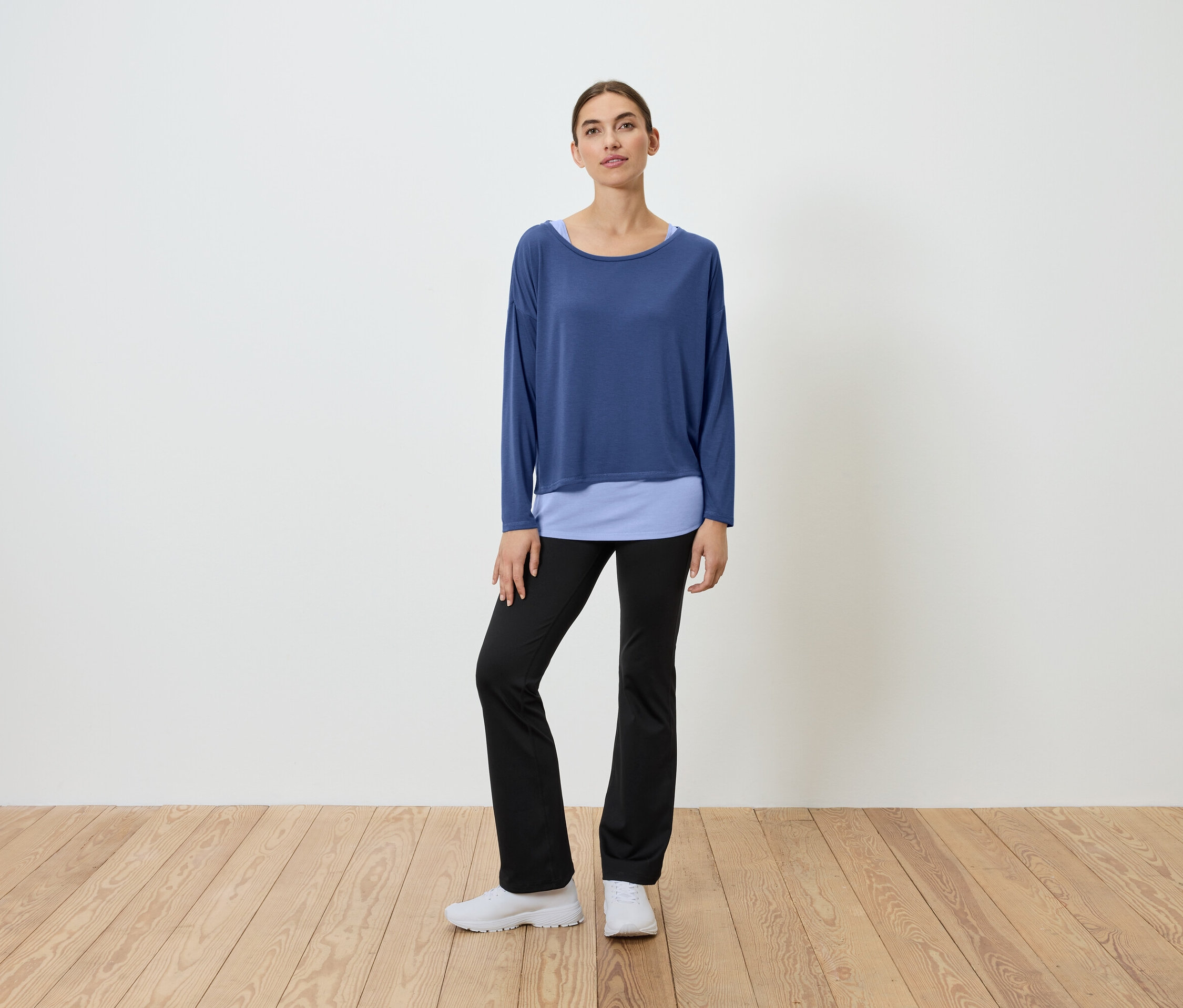 Egy nő áll egy fapadlón, kék felsőt és fekete nadrágot visel.