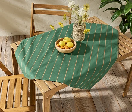 Egy fa asztalon egy kb. 90 x 90 cm-es, csíkos, letörölhető asztalterítő látható. Az asztalterítőn egy gyümölcsös tál és egy váza virágokkal.