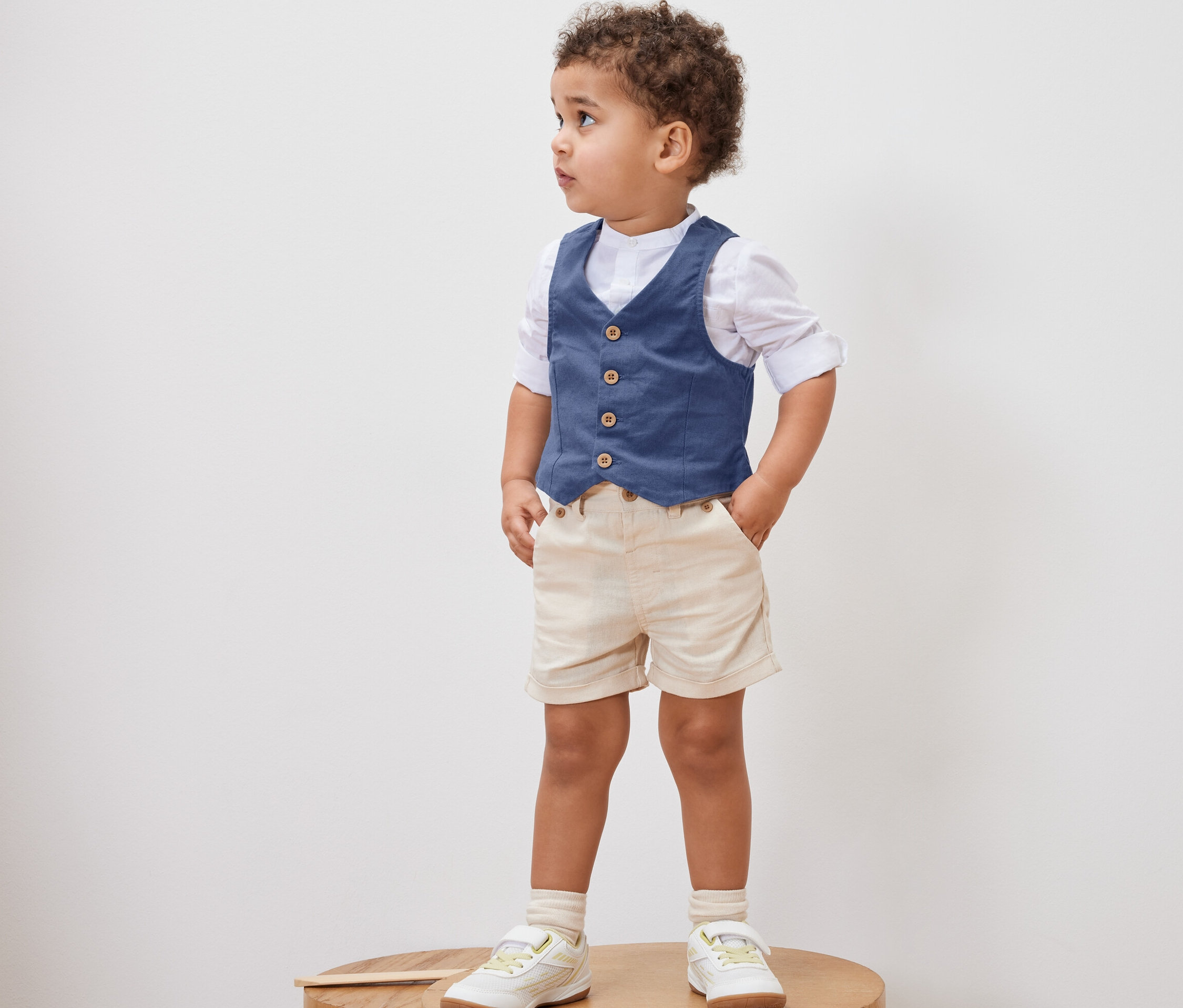 Kisfiú áll egy fa emelvényen, rajta Gyerekmellény, ing, rövidnadrág és cipő.