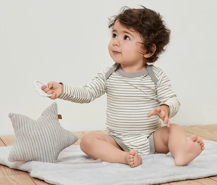 Egy göndör baba ül egy takarón és egy csillaggal játszik. 5 kisgyerek body állítható hosszúsággal van rajta.