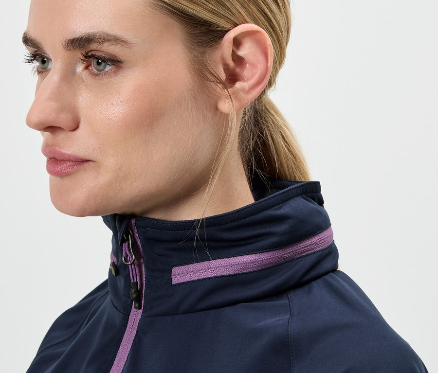 Egy szőke hajú, lófarokba kötött nő arcának közeli képe, aki kék kabátot visel lila részletekkel.