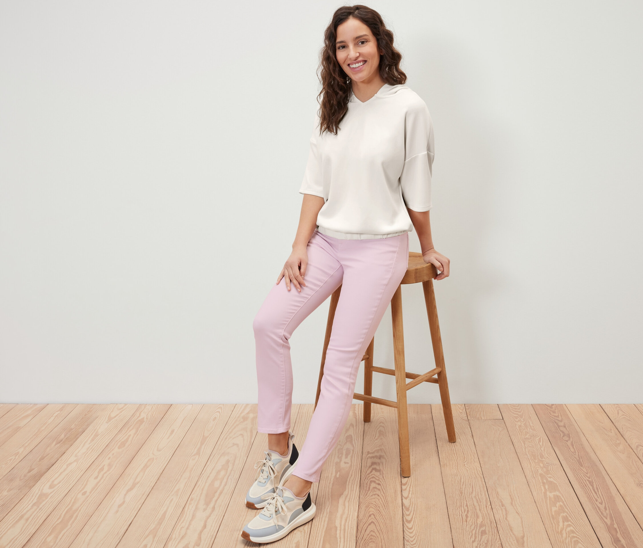 Nő ül egy széken, krémszínű, modálkeverékből készült kapucnis felsőt és rózsaszín nadrágot visel.