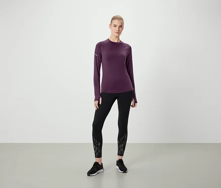 Nő áll sötétlila, hosszú ujjú felsőben, mintás fekete leggingsben és fekete cipőben.