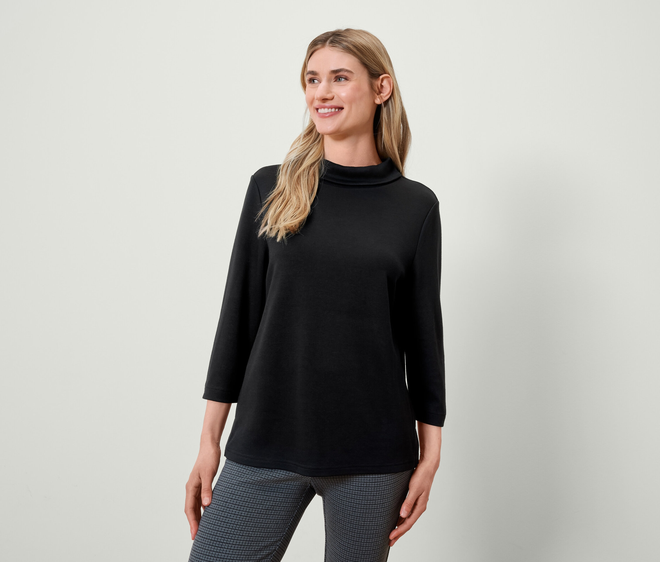 Szőke nő fekete garbópulóverben és szürke mintás nadrágban mosolyog és balra néz.