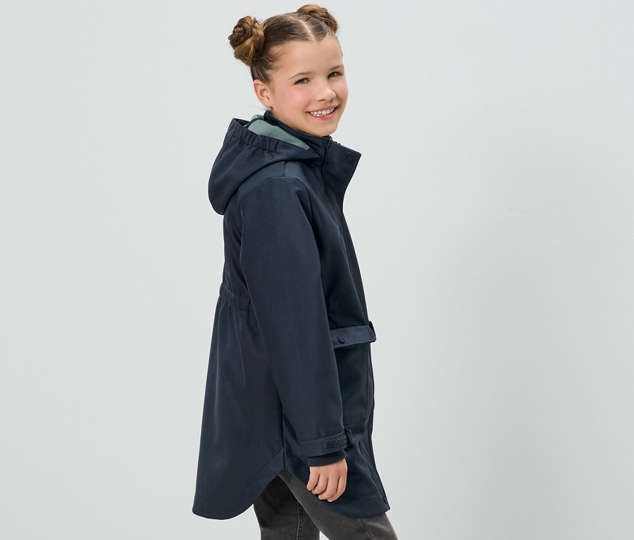 Mosolygó lány pózol egy sötétkék Kinder softshell kabátban és szürke farmerban.
