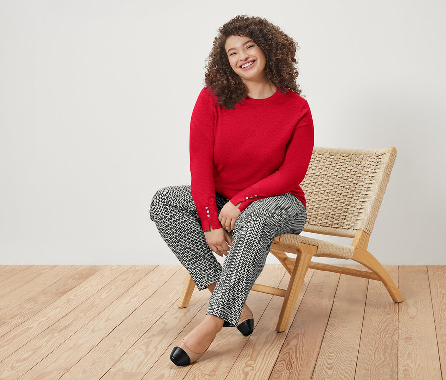 Egy göndör hajú nő ül egy széken, mosolyog, piros pulóvert és mintás nadrágot visel.