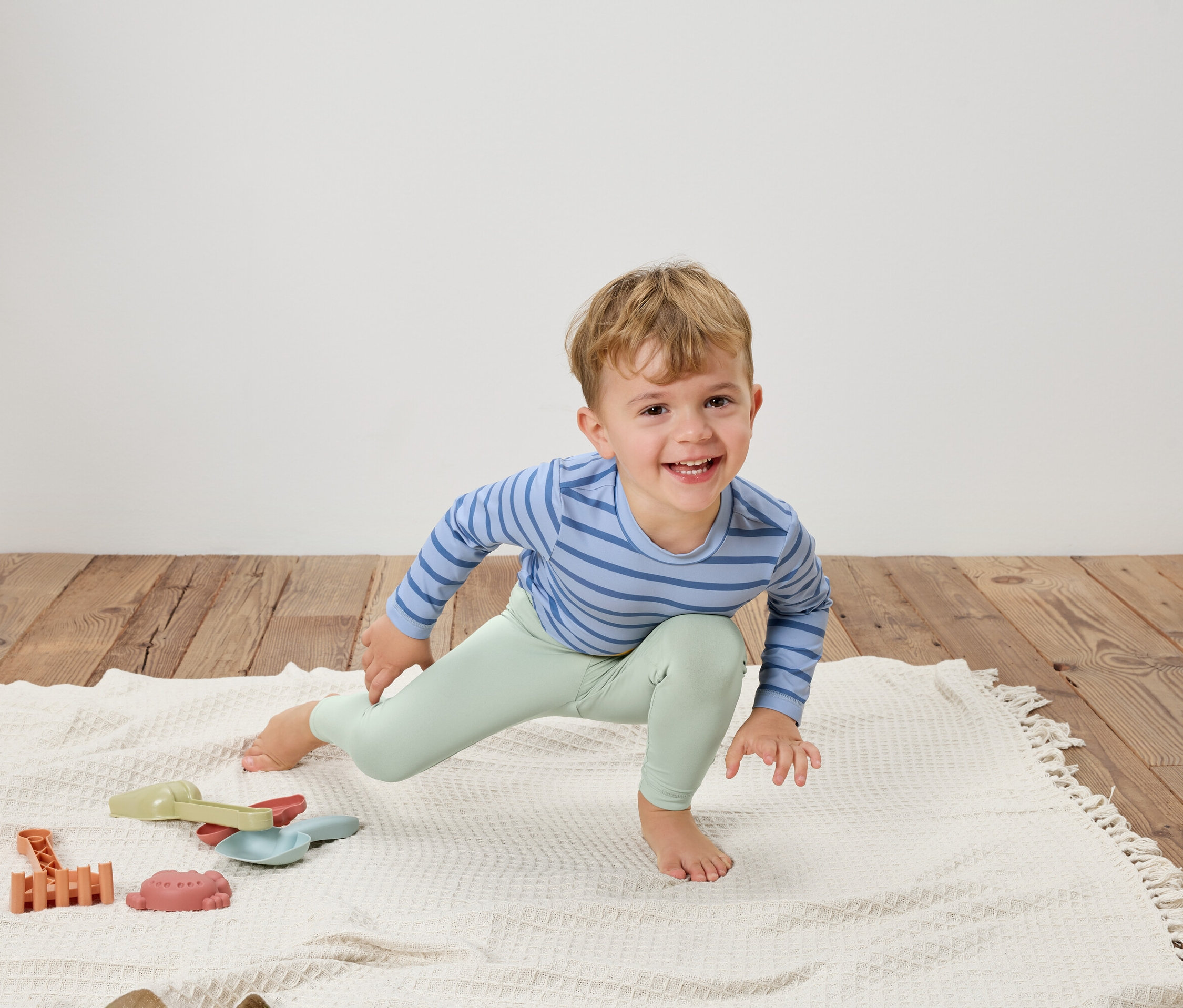 Egy mosolygó kisfiú kék csíkos felsőben és világoszöld leggingsben nyújtózkodik egy fehér takarón játékok mellett.