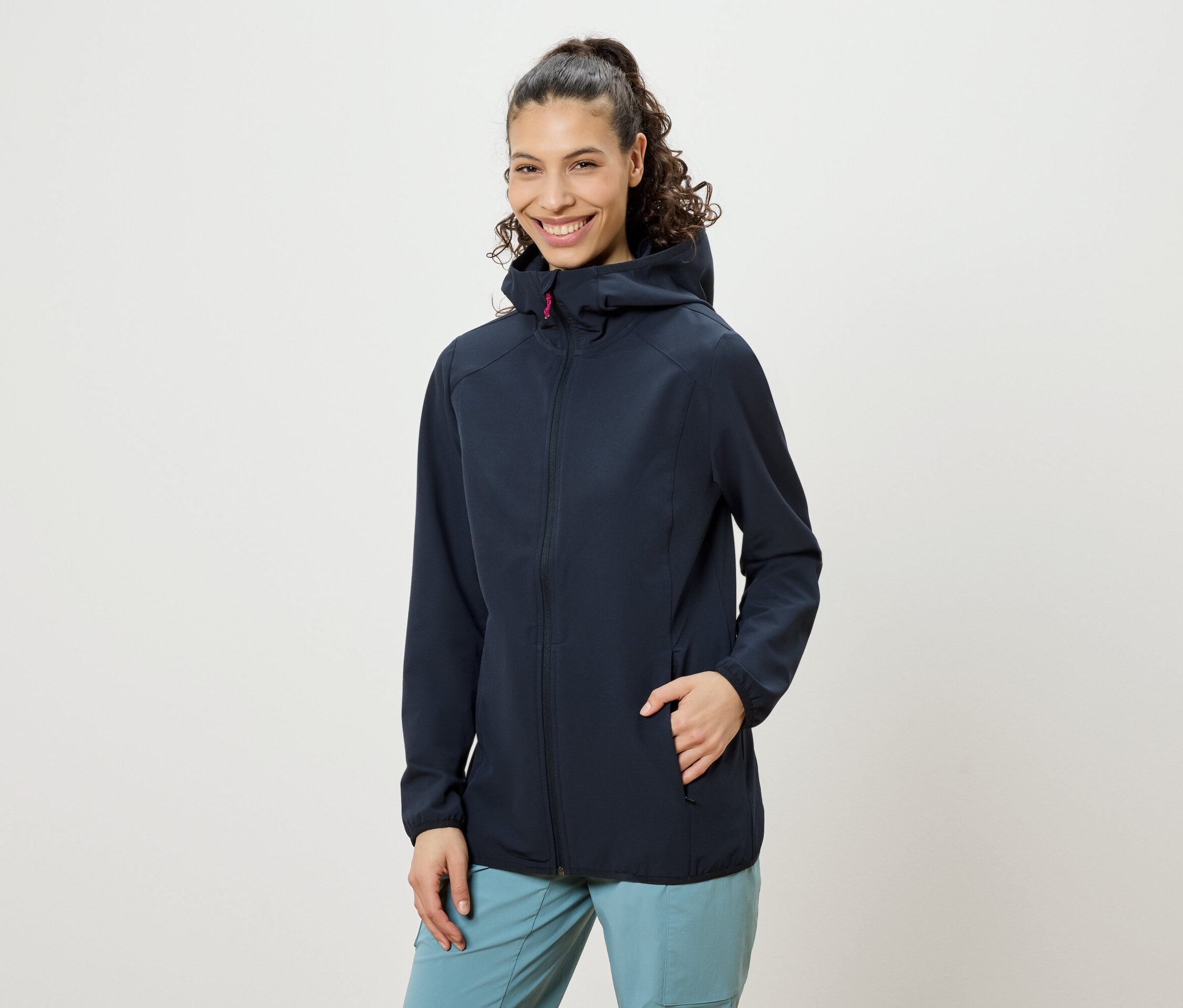 Mosolygó nő, copfba fogott göndör hajjal, sötétkék kapucnis kabátot és kék nadrágot visel, egyik keze a kabátzsebében.