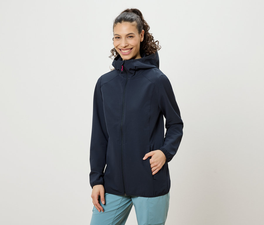 Mosolygó nő, copfba fogott göndör hajjal, sötétkék kapucnis kabátot és kék nadrágot visel, egyik keze a kabátzsebében.