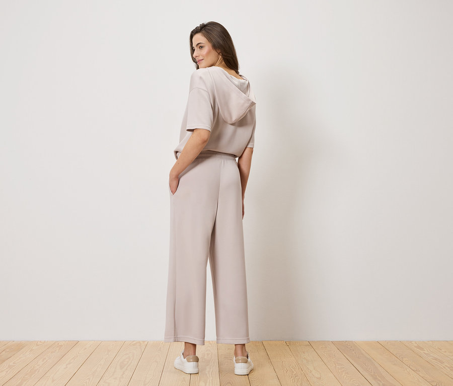 Egy barna hajú nő, hátulról fényképezve, világosbarna kapucnis felsőt és bő nadrágot visel.