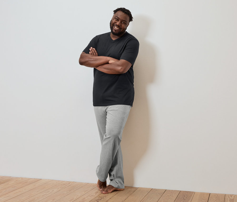 Egy sötét bőrű férfi keresztbe tett karokkal a falnak támaszkodik, fekete pólót és szürke nadrágot visel.