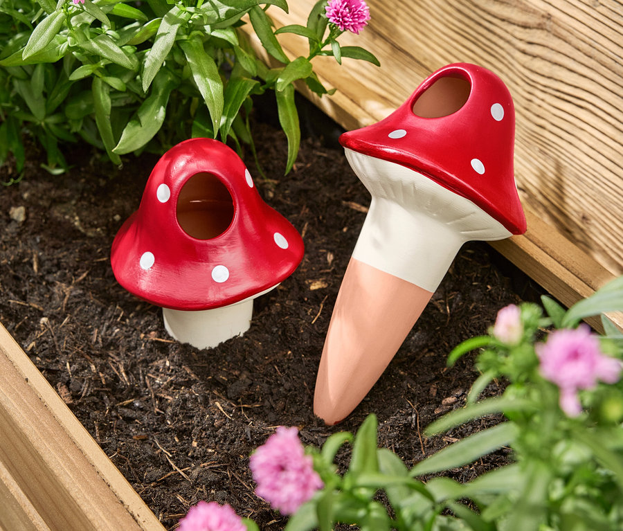 Két piros, gomba alakú, fehér pöttyös öntözőgömb áll egy virágládában virágok és növények között.