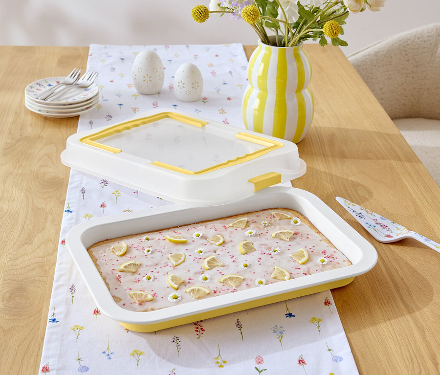 A fa asztalon egy citrommázas és százszorszépekkel díszített sütemény, egy sárga-fehér csíkos kancsó, egy virágmintás tortalapát és egy virágmintás fehér asztalterítő található.