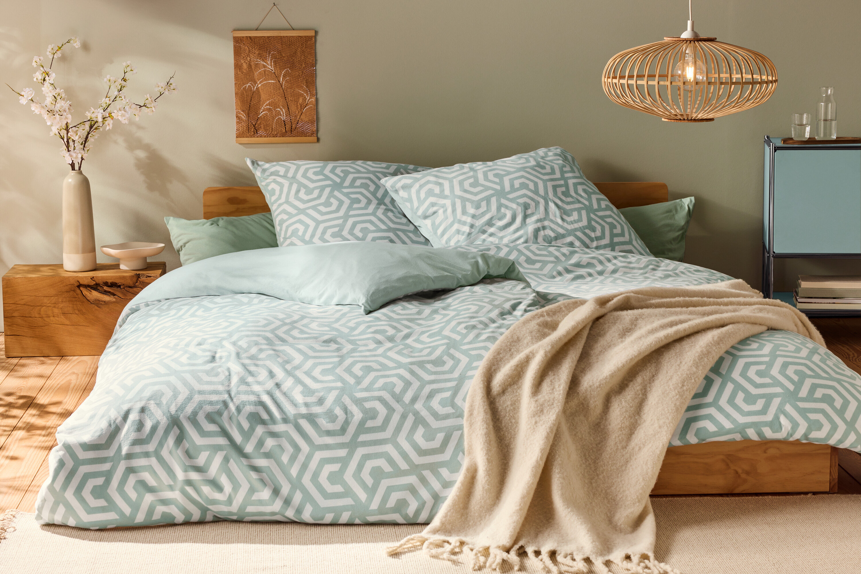 Ágy normál méretű, geometrikus mintázatú dzsörzé ágyneművel és világoszöld párnákkal.
