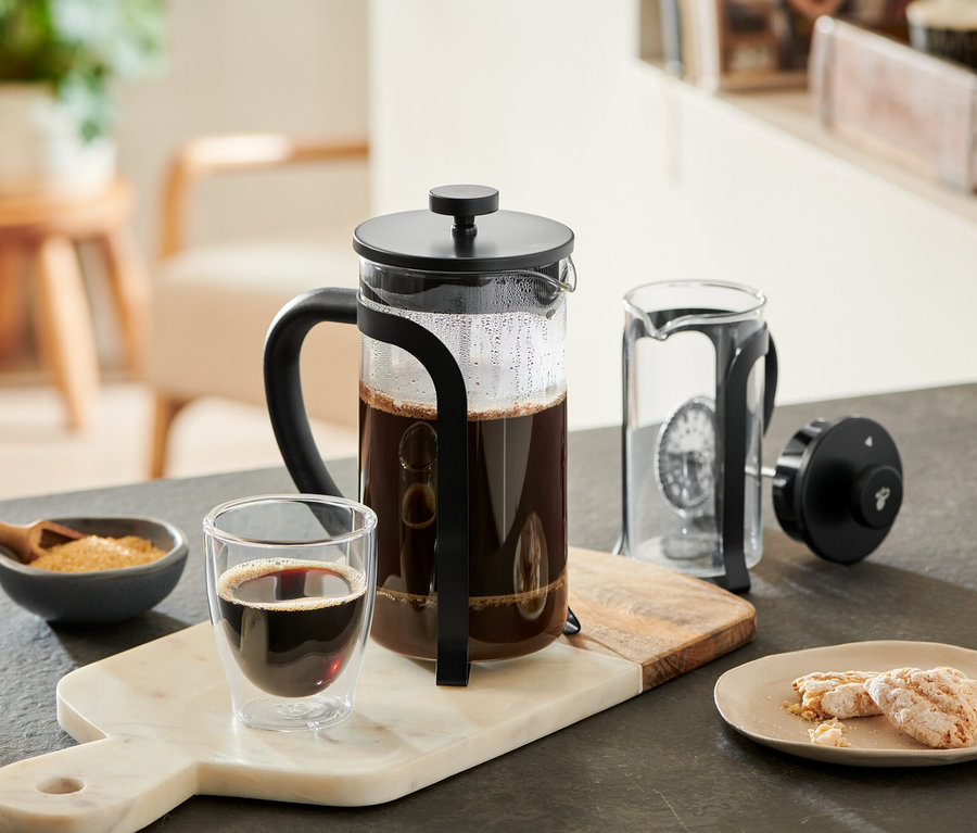 Egy fekete dugattyús kávéfőző 800 ml, 6 csésze, kávéval töltve, márvány deszkán áll egy pohár kávé, egy cukortartó tál és sütemények mellett. Egy másik dugattyús kávéfőző szétszerelve fekszik mellette.