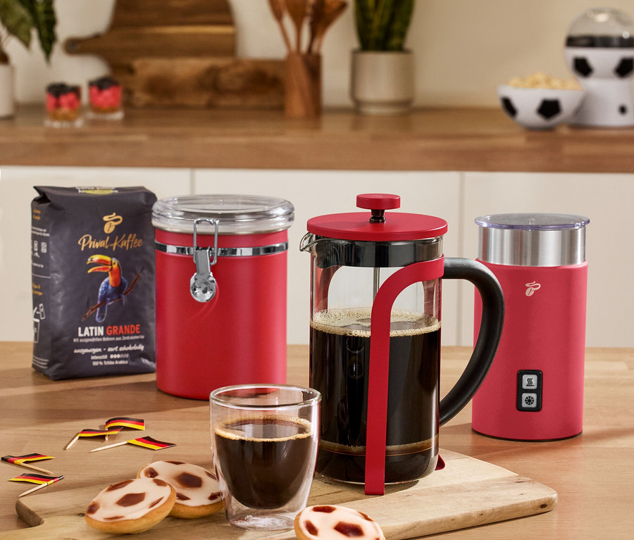 Az asztalon piros aromatartó doboz, piros elektromos tejhabosító, piros dugattyús kávéfőző, 2 duplafalú caffè crema pohár és egy csomag kávé látható.