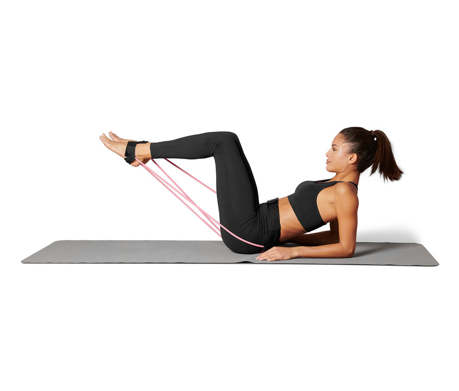 Nő fekszik a matracon, és a lábához rögzített derékra rögzíthető fitnesz gumikötéllel edz.
