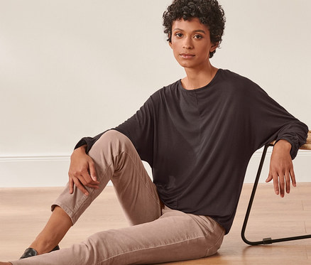 Nő ül a padlón egy fonott szék mellett, sötétbarna denevérujjú felsőben.