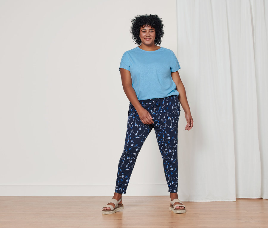 Nő áll világoskék lenvászon pólóban, sötétkék virágos dzsörzénadrágban és bézs bőr szandálban.