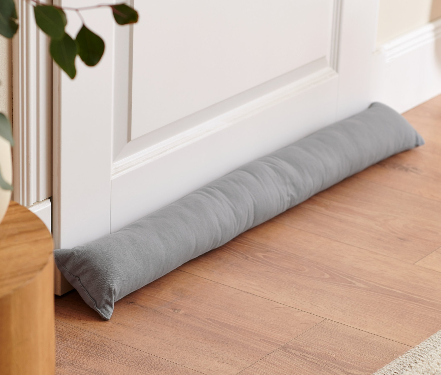 Szürke huzatfogó párna fekszik a fehér ajtó alján a fapadlón.