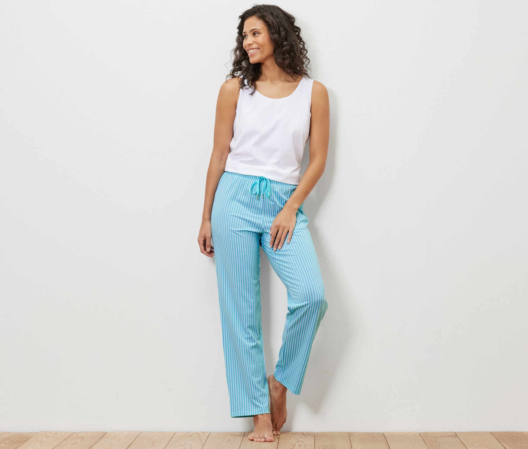 A falnak támaszkodó nő fehér trikót és kék csíkos pizsamanadrágot visel.