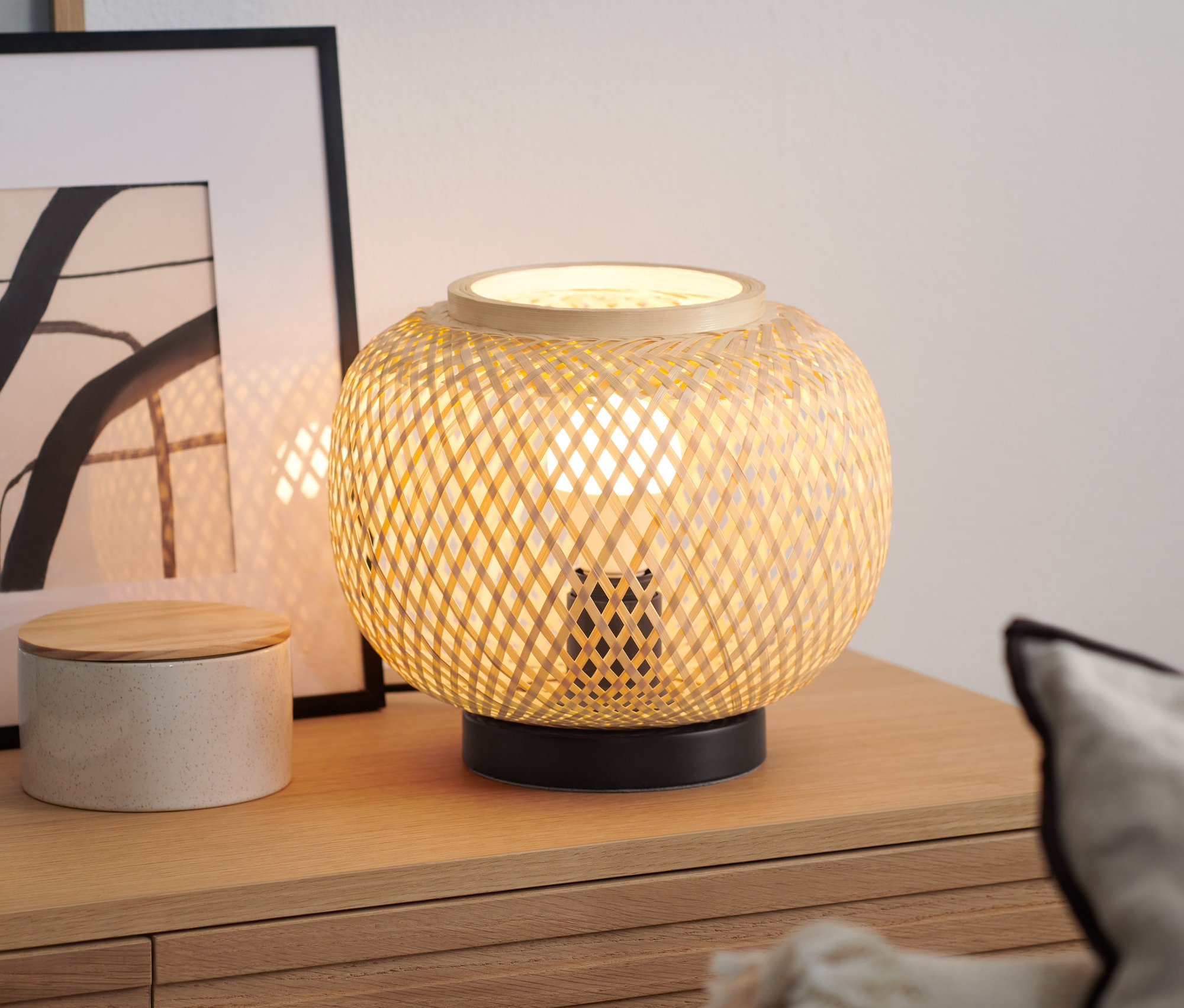 Bambusz lámpaernyős asztali lámpa áll egy fa komódon egy könyv és egy dekoratív doboz mellett.