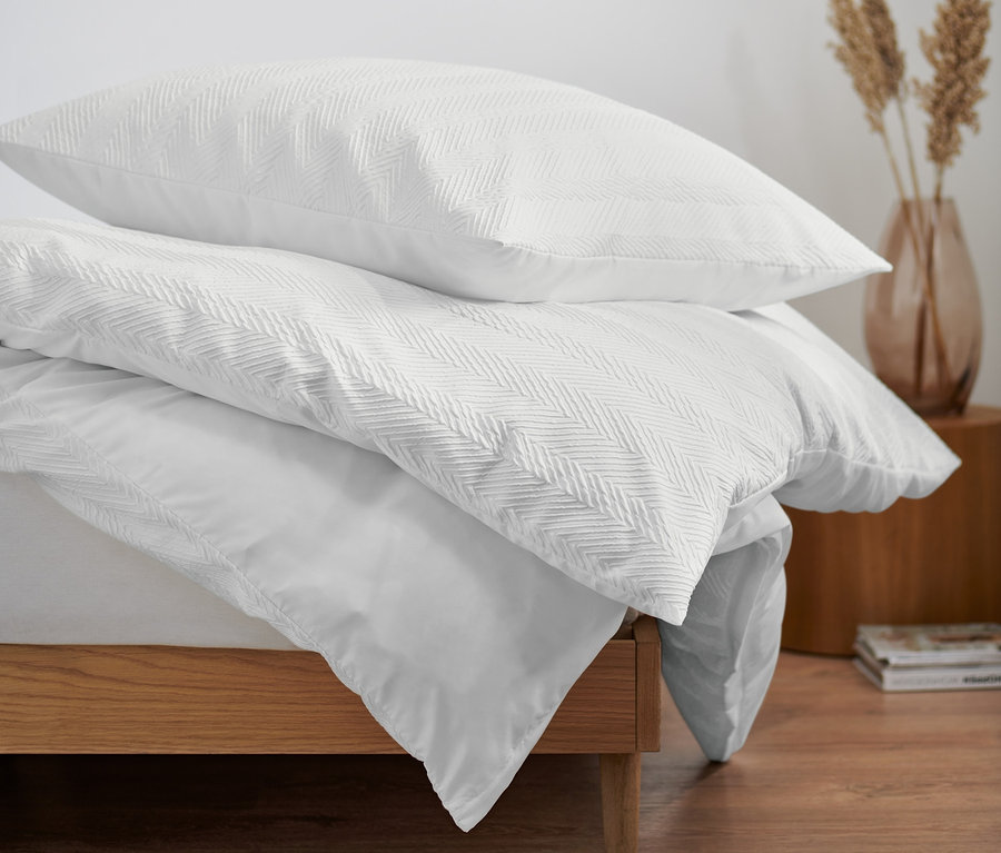 Három darab strukturált ágynemű egyszemélyes méretben egy ágyon. A háttérben egy kerek tükör és egy váza látható.