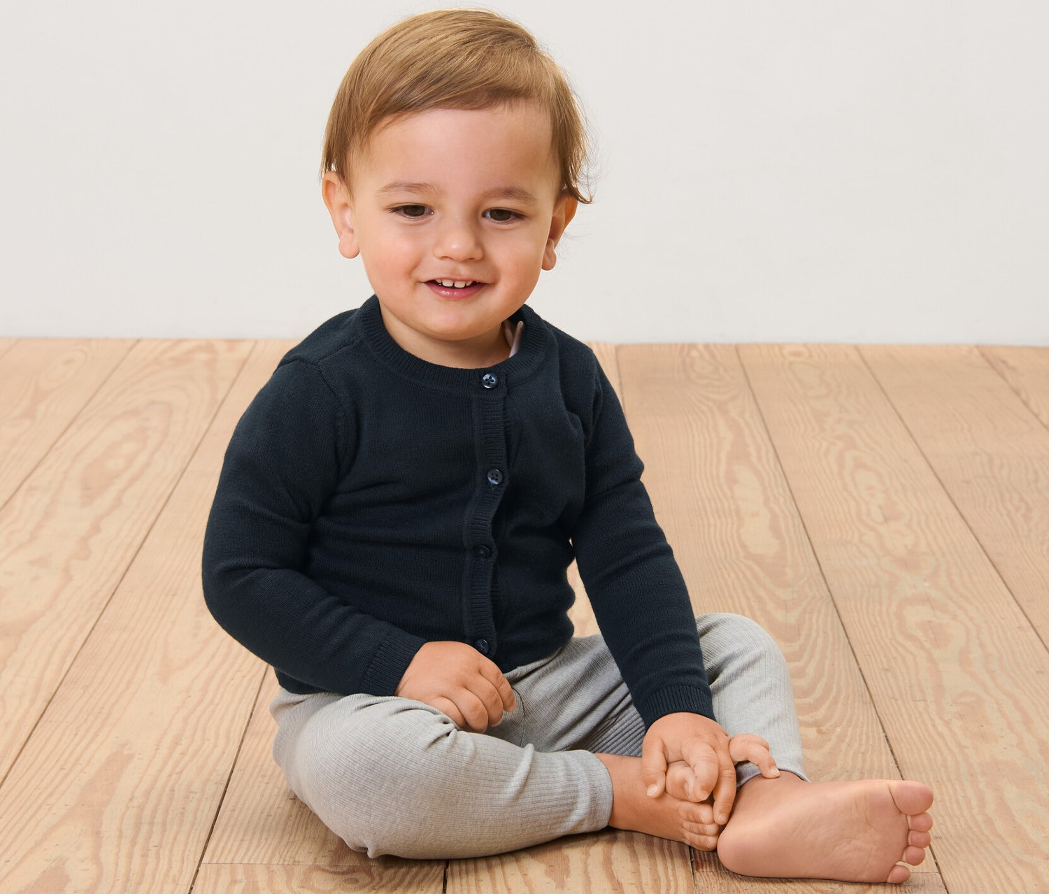 Egy gyermek ül egy fapadlón, kötött gyerekkardigánt visel.