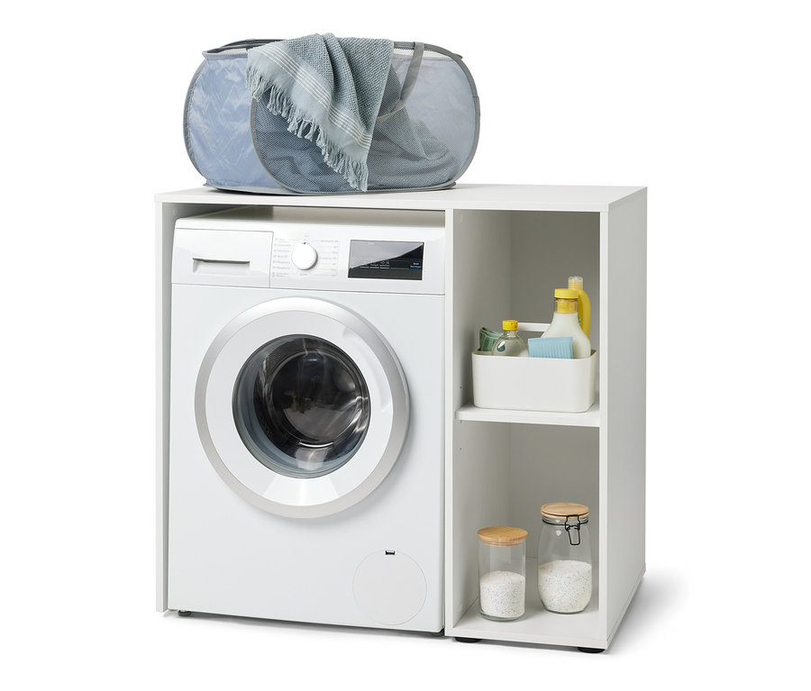 Egy fehér mosógép alatt egy fehér mosógép szekrény látható oldalsó tárolóval. A szekrényen egy szürke szennyestartó van.