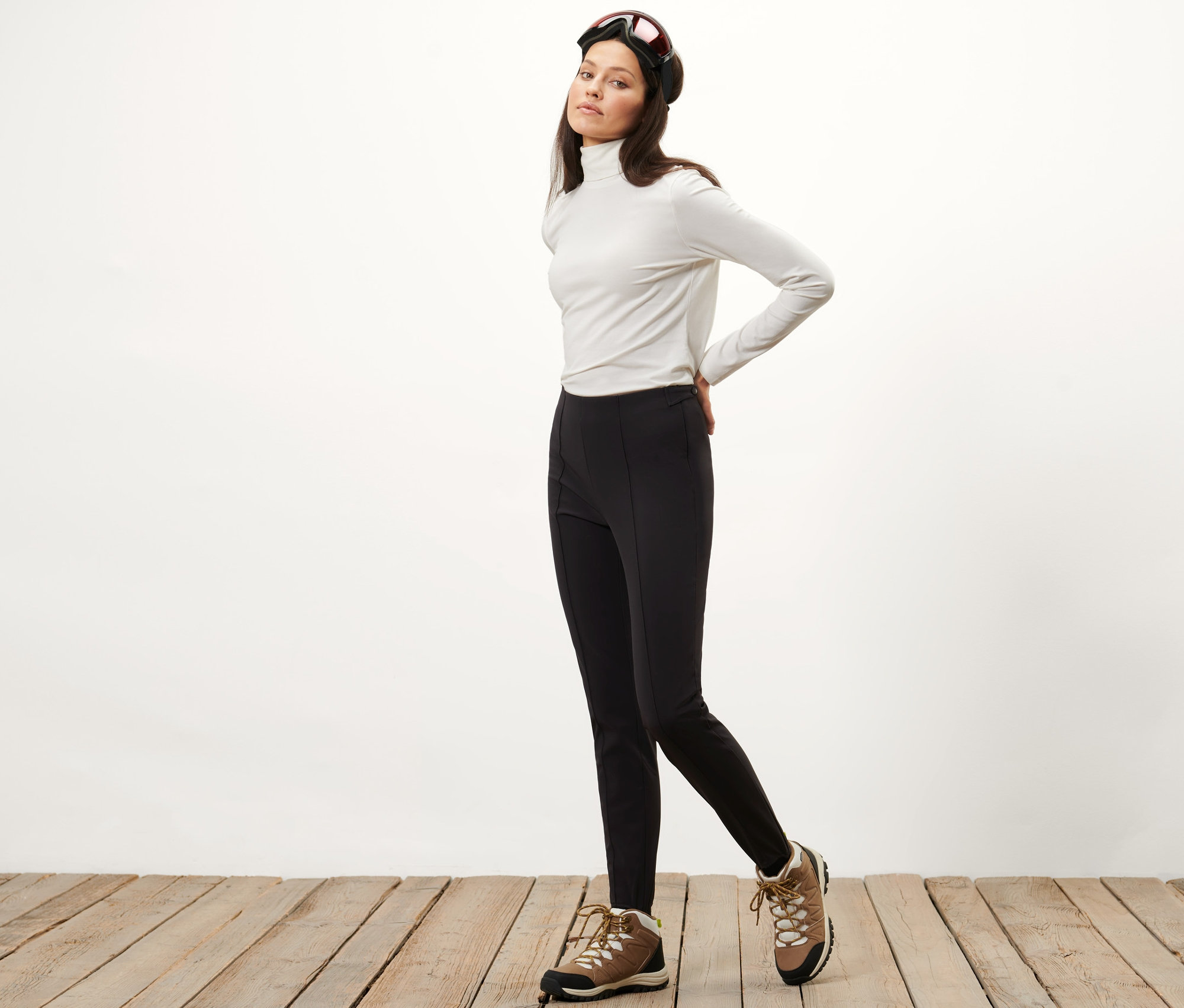 Nő pózol fekete női softshell sínadrágban, fehér garbóban és sí szemüvegben.