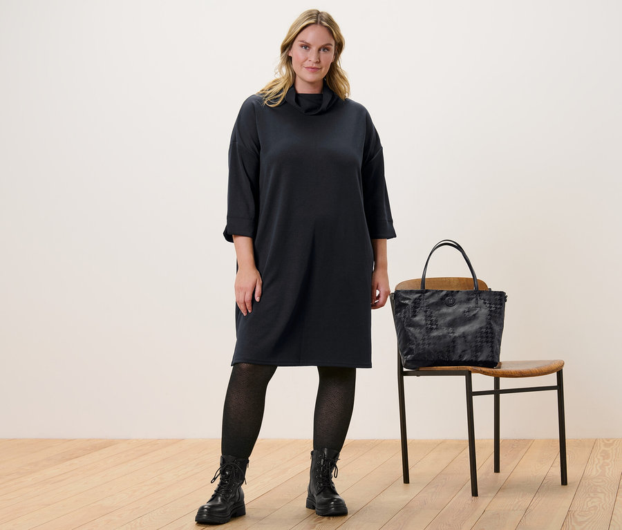 Egy nő áll, és egy fekete női 3/4-es ujjú lezser ruhát és egy antracit női strukturált harisnyát visel. Mellette egy szék áll, rajta egy kézitáska.