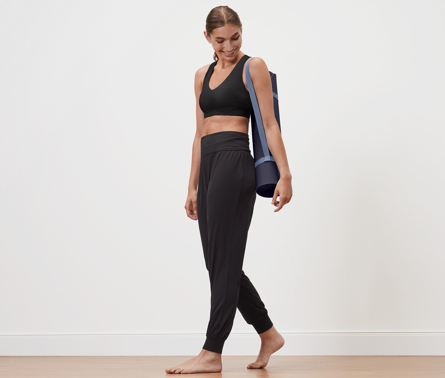 Nő sétál, jógamatracot visz a vállán. Fekete női jóganadrágot visel.